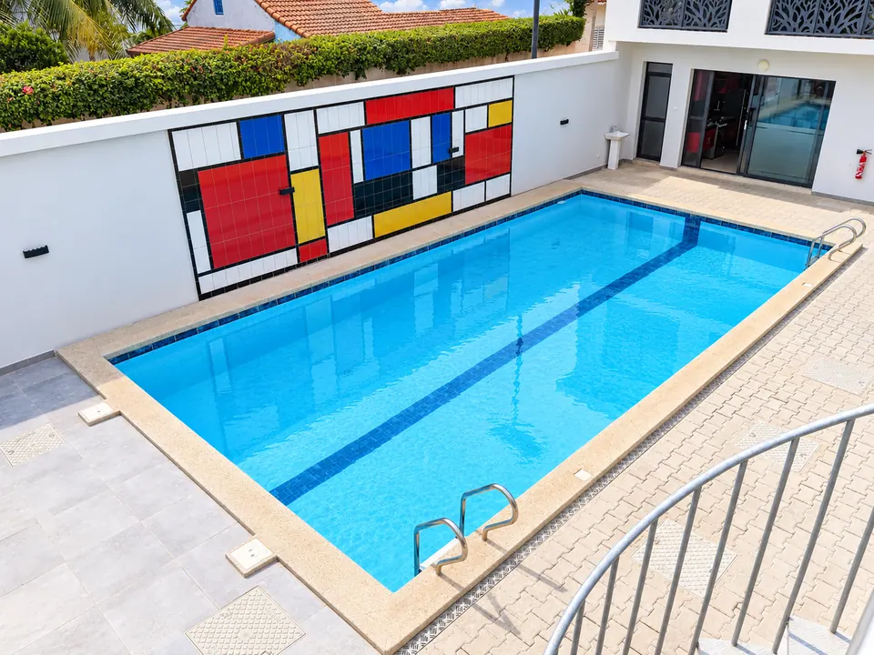Vue plongeante sur la piscine de la résidence, bordée d'un mur orné d'une fresque en carreaux rouges, bleus, jaunes et blancs aux lignes noires