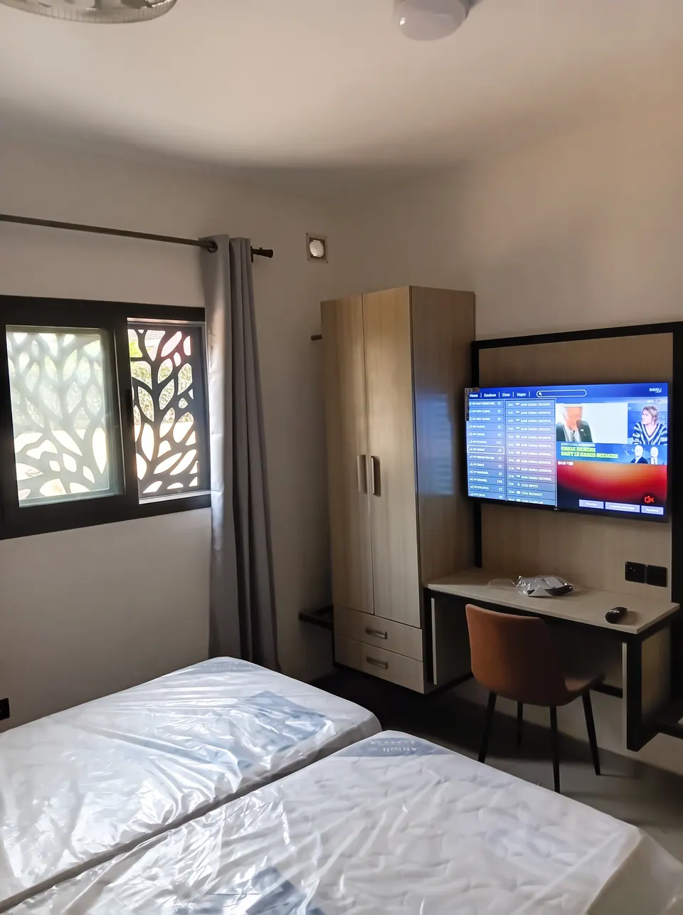 Chambre avec deux lits, une armoire, un bureau, un téléviseur mural et une fenêtre à motif ajouré